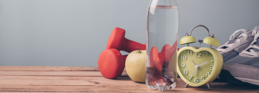 Dumbells, water bottle, apple, workout shoes, and alarm clock placed on wood table